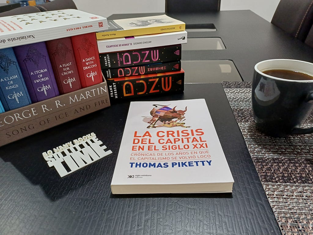 Books on a dark table beside a mug of coffee: Thomas Piketty’s “La crisis del capital en el siglo XXI”, the Dune saga, Marcus Aurelius’s Meditations, “Descartes’ Error” and A Song of Ice and Fire, next to a sign reading “So many books, so little time”